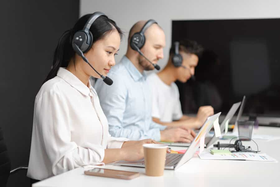 hree people working in a call center sitting at a desk with headset on and with their laptops